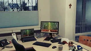 Screibtisch im Pfarrbüro ausgestattet mit vielen Dingen: Bildschirm und Tastatur, Laptop, Telefon ...