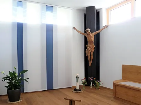 Kleiner Gebetsraum mit schlichtem Holzkreuz an einer dunklen Wand, davor Kerzen und Blumen. 