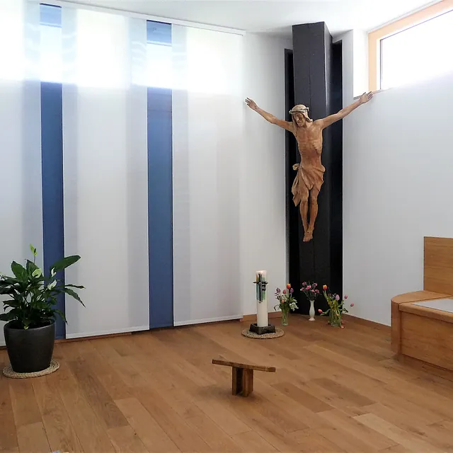Kleiner Gebetsraum mit schlichtem Holzkreuz an einer dunklen Wand, davor Kerzen und Blumen. 