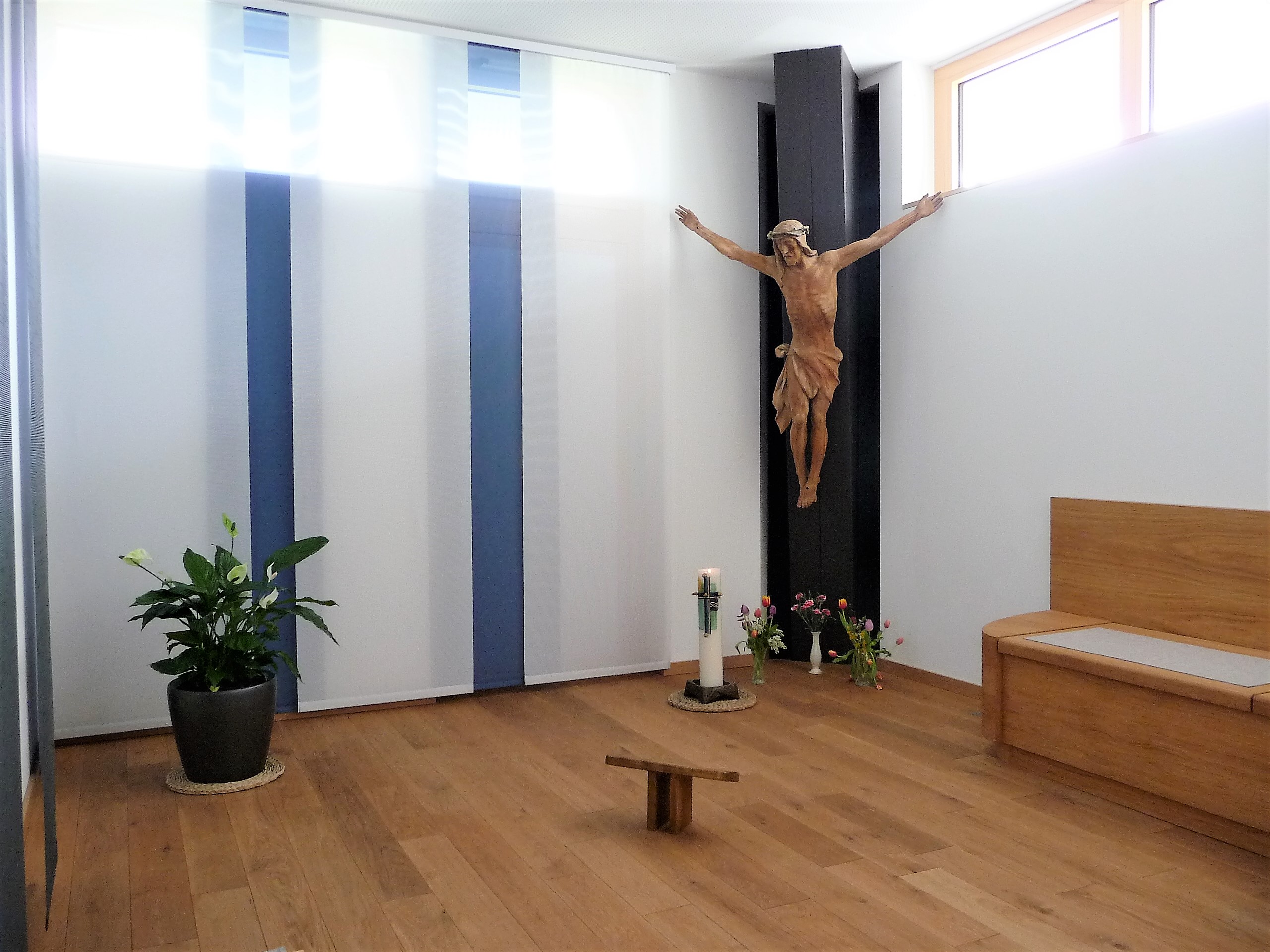 Kleiner Gebetsraum mit schlichtem Holzkreuz an einer dunklen Wand, davor Kerzen und Blumen. 
